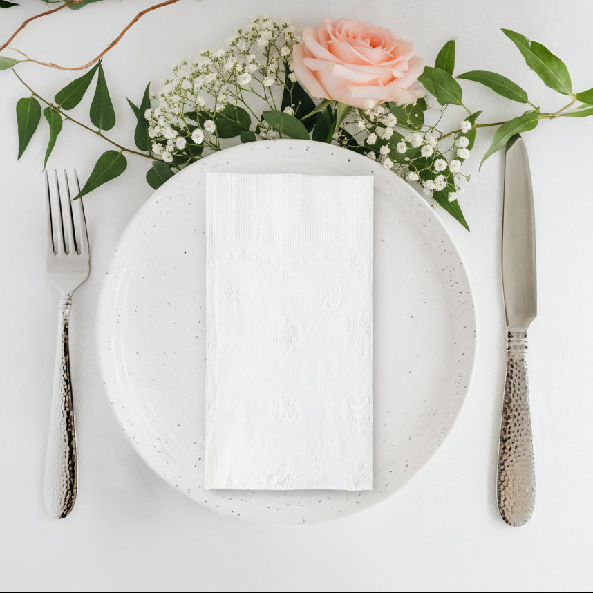 White plate with a folded white napkin, silver fork, and knife on a light gray background with greenery and flowers.